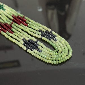Puede incluir: Un collar de múltiples hilos con pequeñas cuentas facetadas en tonos verde claro, verde oscuro, rojo oscuro y negro. El collar está dispuesto en un patrón en cascada, mostrando la variedad de colores y texturas.