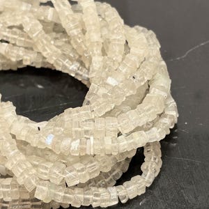 Puede incluir: Primer plano de un montón de cuentas translúcidas, de corte cuadrado. Las cuentas son de un color blanco roto pálido y parecen estar hechas de un material transparente, posiblemente cristalino. Las cuentas están ensartadas, formando un hilo flexible, para joyería.