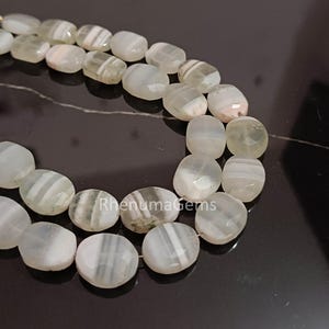May include: Close-up of two strands of gemstone beads. The beads are oval-shaped with a mix of white, gray, and translucent sections. The beads have a polished surface and are strung together. The background is a dark, reflective surface.