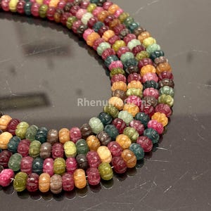 Può includere: Un filo di perline di tormalina multicolore, ognuna caratterizzata da una forma intagliata, scanalata e simile a una zucca. Le perline mostrano una sfumatura di tonalità rosa, verde, gialla e marrone. Il testo Rhenu Gems è visibile al centro.