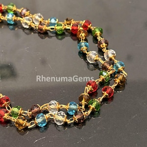 Puede incluir: Un collar de varias hebras con cuentas de cristal facetado en colores rojo, verde, azul, marrón y transparente, unidas con alambre de metal dorado. El texto RhenumaGems es visible en el centro de la imagen, integrado en la presentación.