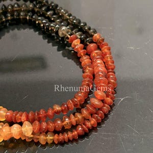 Pode incluir: Um colar de várias camadas com pequenas contas em forma de disco num gradiente de cores, que transita de cinzento escuro translúcido para laranja vibrante e amarelo claro. O texto RhenumaGems é visível no centro das camadas de contas.