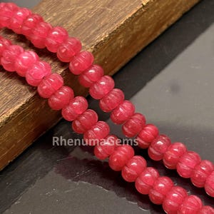 May include: A strand of vibrant red, carved, round beads with a ribbed texture. The beads are arranged in two parallel lines, showcasing a consistent shape and deep crimson color. The text RhenumaGems is visible in white lettering across the beads.