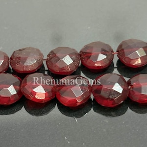 Peut inclure: Gros plan sur un fil de perles de pierres précieuses rouge foncé à facettes. Les perles sont d'une couleur riche et translucide, avec de multiples facettes captant la lumière. Les perles sont enfilées. Le texte "Rhenuma Gems" est visible.