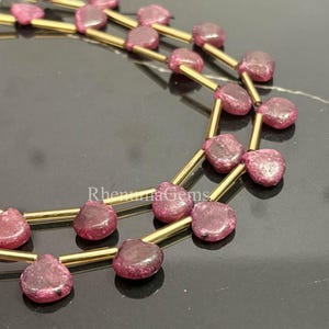 Könnte beinhalten: Nahaufnahme von zwei Strängen herzförmiger, tiefrosa Edelsteine, die auf goldfarbenen Metallstäben aufgereiht sind. Die Edelsteine haben eine leicht raue Textur. Der Hintergrund ist eine dunkle, reflektierende Oberfläche. Der Text "Rhenuma Gems" ist sichtbar.