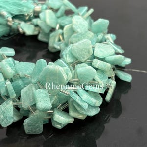 Peut inclure: Gros plan sur un fil de perles de pierres précieuses amazonite bleu clair. Les perles sont de forme irrégulière, avec une surface polie. Le fil est enfilé avec du fil argenté. Un pompon assorti est visible en arrière-plan. Le texte "RhenumaGems" est visible.