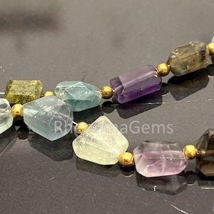 May include: A beaded necklace featuring irregular, faceted gemstones in shades of purple, green, blue, and brown, interspersed with small, round, metallic gold-coloured beads. The text RhoaGems is visible in the centre of the image.