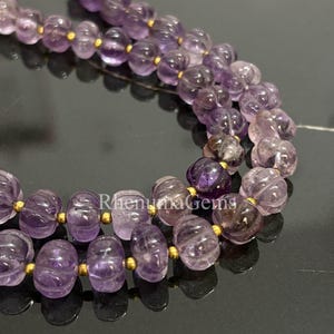 Könnte beinhalten: Nahaufnahme von zwei Strängen Amethyst-Edelsteinperlen. Die Perlen sind mit einem gerippten Design geschnitzt und mit goldfarbenen Abstandshaltern verbunden. Die Amethystfarbe reicht von hell- bis tiefviolett.