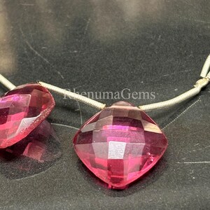 Könnte beinhalten: Zwei facettierte, kissenförmige, tiefrosa Edelsteine, die auf einen dünnen, silberfarbenen Draht aufgezogen sind. Die Steine sind durchscheinend mit einer geometrischen, facettierten Oberfläche, die Licht reflektiert. Der Text "RhenumaGems" ist sichtbar.