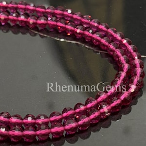 Könnte beinhalten: Nahaufnahme von drei Strängen facettierter, tiefmagentafarbener Edelsteine. Die Perlen sind zu einer Halskette oder einem Armband aufgereiht. Der Text "RhenumaGems" ist im unteren Zentrum des Bildes zu sehen. Die Edelsteine haben ein funkelndes, poliertes Aussehen.