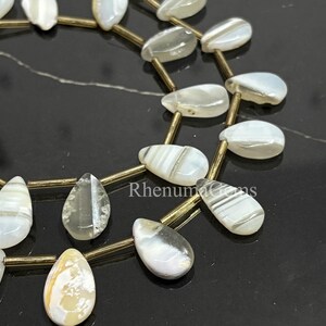 Könnte beinhalten: Nahaufnahme einer Kette aus perlmuttartigen, tropfenförmigen Perlen. Die Perlen sind auf goldfarbenem Draht aufgefädelt und haben ein subtiles, gestreiftes Muster. Die Perlen sind weiß, cremefarben und grau.