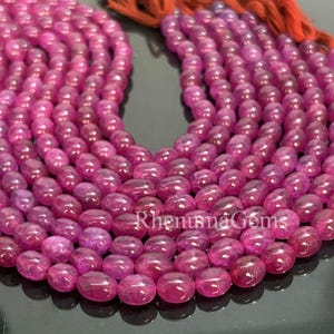 Könnte beinhalten: Stränge aus polierten, oval geformten Rubinperlen in verschiedenen Rosa- und Rottönen. Die Perlen sind aufgefädelt, mit einer Quaste aus rotem Faden oben. Die Perlen sind glatt und glänzend.