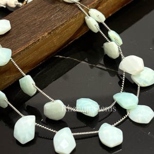 Könnte beinhalten: Stränge aus hellblauen und weißen Edelsteinperlen. Die Perlen sind facettiert und auf einem Silberdraht aufgefädelt. Die Edelsteine haben eine unregelmäßige Form und werden auf einer dunklen Oberfläche präsentiert.