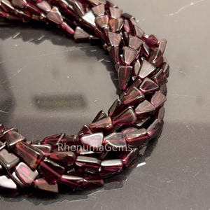 Puede incluir: Primer plano de un collar de cuentas triangulares de color rojo oscuro. Las cuentas están ensartadas, formando una forma curva. Las cuentas tienen un acabado brillante y están dispuestas en un patrón en capas. El texto "RhenumaGems" es visible.
