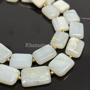 May include: Close-up of a necklace featuring rectangular, off-white gemstones with gold-colored accents. The gemstones are strung together, creating a delicate and elegant piece of jewelry. The necklace is displayed on a dark surface.