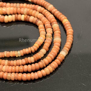 May include: A strand of smooth, round, peach-colored beads arranged in a circular pattern. The beads exhibit subtle variations in tone, ranging from light cream to deep salmon, with a polished finish. The text RhenumGems is visible in white.