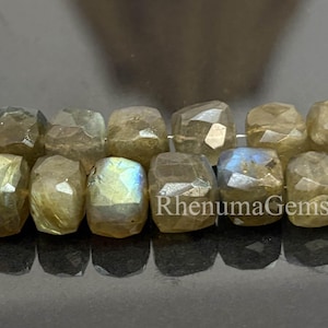 Könnte beinhalten: Eine Kette aus facettierten Labradorit-Perlen. Die Perlen sind eine Mischung aus Grau, Beige und irisierendem Blaugrün. Die Perlen sind würfelförmig und haben eine polierte Oberfläche. Der Text "RhenumaGems" ist sichtbar.