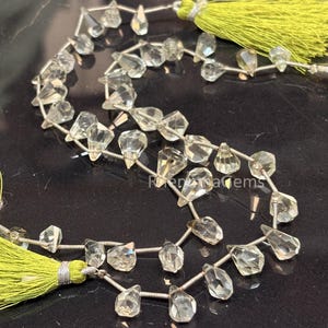May include: A strand of faceted, clear, pear-shaped quartz beads strung on a silver-toned wire, featuring a bright lime green tassel at each end. The beads are arranged in a curved line, showcasing their transparent, multi-faceted surfaces.