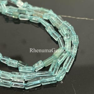 Könnte beinhalten: Nahaufnahme von Strängen hellblauer, rechteckiger Edelsteinperlen. Die Perlen sind durchscheinend und haben eine leicht unregelmäßige Form. Der Text "RhenumaGems" ist im Bild zu sehen. Diese Perlen eignen sich für die Schmuckherstellung.
