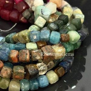 Puede incluir: Primer plano de una colección de cuentas de piedras preciosas en varios colores, incluyendo azul, verde, marrón y rosa. Las cuentas están facetadas y tienen forma de cubo. El texto "RhenumaGems" es visible.