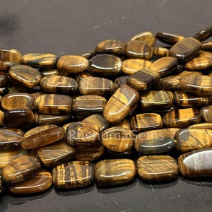 Könnte beinhalten: Nahaufnahme einer Sammlung polierter Tigeraugen-Edelsteinperlen. Die Perlen sind rechteckig mit abgerundeten Kanten und zeigen eine Mischung aus braunen, goldenen und schwarzen Farben. Die Edelsteine haben ein natürliches, gestreiftes Muster.