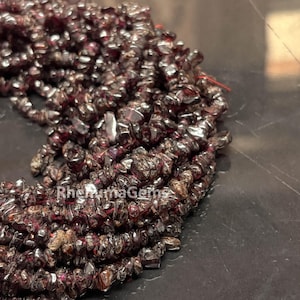 Può includere: Una collezione di piccole schegge irregolari di pietra preziosa granato rosso scuro, infilate insieme. Le superfici scure e levigate delle pietre riflettono la luce e il testo RhenumaGems è visibile in bianco in basso a sinistra.