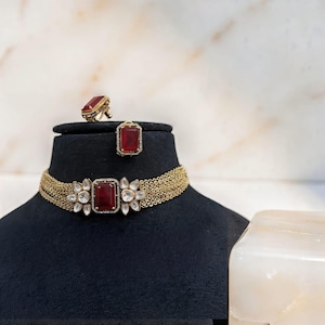 May include: A gold-coloured choker necklace, ring, and earrings set. The choker features multiple strands and a large, rectangular, red gemstone. The ring and earrings also have red gemstones. The jewellery is displayed on a black mannequin.