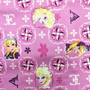 May include: Pink fabric with a repeating pattern featuring cartoon character portraits, floral designs, and decorative elements. The design includes portraits of a blonde character with blue eyes, set against a background of white and dark pink shapes.