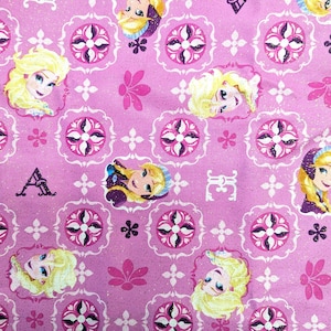 May include: Pink fabric featuring a repeating pattern of a cartoon character's face, floral designs, and decorative elements. The character has blonde hair and is framed by a decorative border. The background is a solid pink color.