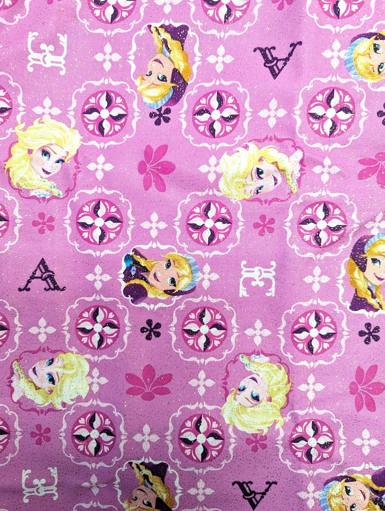 May include: Pink fabric featuring a repeating pattern of a cartoon character's face, floral designs, and decorative elements. The character has blonde hair and is framed by a decorative border. The background is a solid pink color.