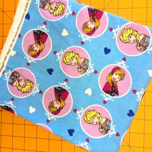 May include: Light blue fabric featuring circular portraits of two cartoon characters in pink circles, with white decorative swirls, white and blue hearts. The fabric has text along the edge that reads "5. Sold for noncommercial home use only. Other uses prohibited Do not use for".