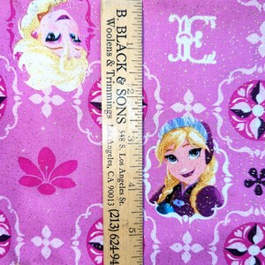 May include: Pink fabric with a repeating pattern of cartoon characters and floral designs. The fabric has a glittery texture and includes the text "B. BLACK & SONS" and a phone number on a wooden ruler.