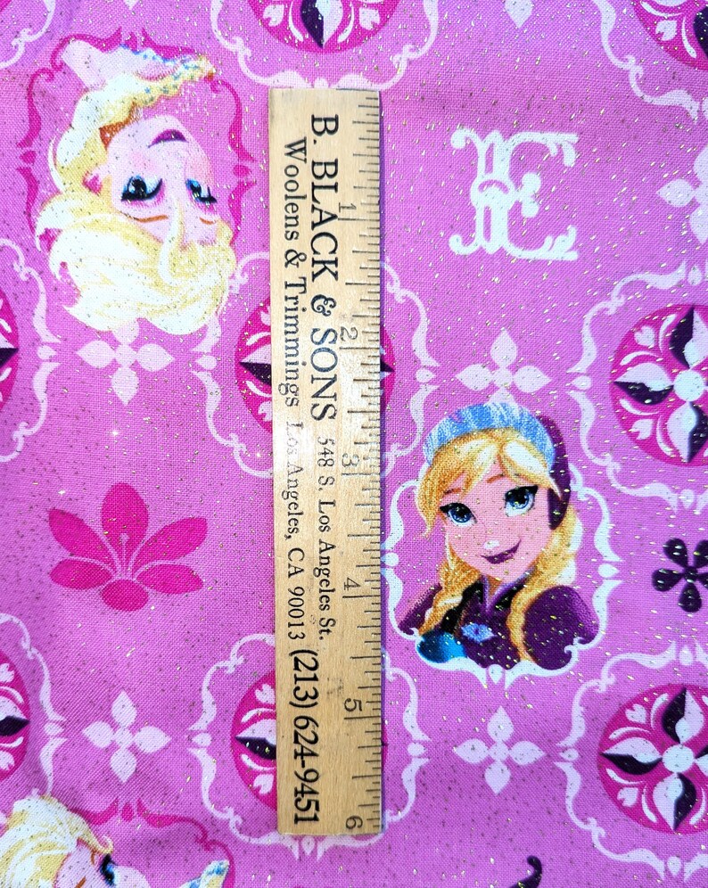 May include: Pink fabric with a repeating pattern of cartoon characters and floral designs. The fabric has a glittery texture and includes the text "B. BLACK & SONS" and a phone number on a wooden ruler.