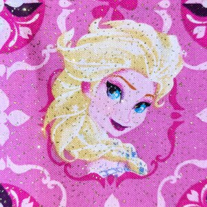 May include: A close-up of a pink fabric featuring a portrait of a blonde woman with blue eyes, likely a character from a popular animated film. The background has a repeating pattern of pink and white floral designs with gold glitter accents.