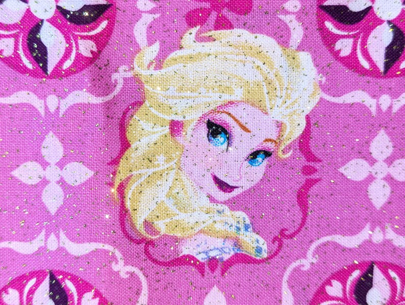 May include: A close-up of a pink fabric featuring a portrait of a blonde woman with blue eyes, likely a character from a popular animated film. The background has a repeating pattern of pink and white floral designs with gold glitter accents.