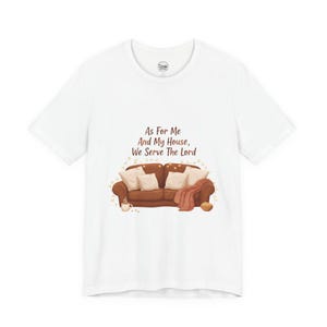 May include: White t-shirt featuring a graphic of a brown sofa with pillows, a blanket, and a coffee cup. The text "As For Me And My House, We Serve The Lord" is printed above the sofa. The design has a cozy, homey feel.