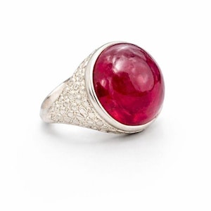 May include: A silver ring featuring a large, vibrant red gemstone. The gemstone is smooth and rounded, set in a bezel. The band of the ring has a textured, patterned design. The ring is displayed against a white background.