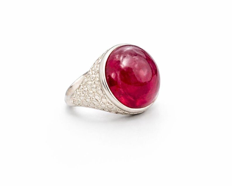 Może przedstawiać: Srebrny pierścionek z dużym, żywym, czerwonym kamieniem szlachetnym. Kamień szlachetny jest gładki i zaokrąglony, osadzony w oprawie. Obrączka pierścionka ma wzorzystą fakturę. Pierścionek jest prezentowany na białym tle.