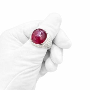 Może przedstawiać: Duży, srebrny pierścionek z wyraźnym, okrągłym, głęboko czerwonym kamieniem szlachetnym. Opaska pierścionka ma teksturowany, tkany wz&oacute;r. Pierścionek jest trzymany przez dłoń w rękawicy na białym tle, podkreślając biżuterię.