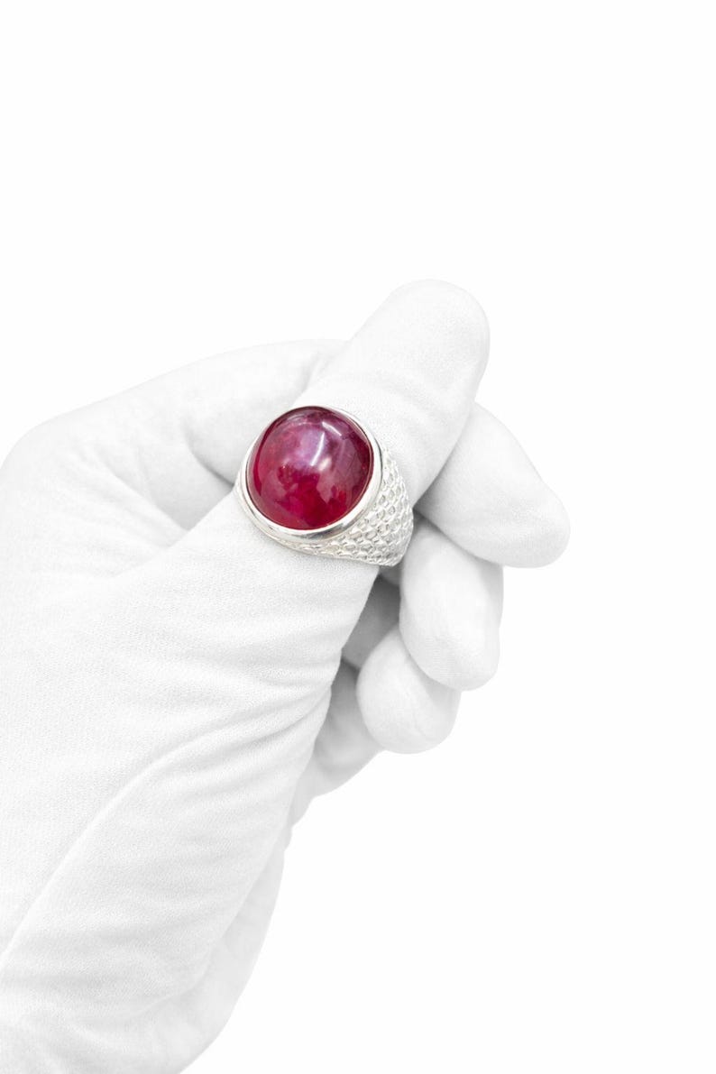 Może przedstawiać: Duży, srebrny pierścionek z wyraźnym, okrągłym, głęboko czerwonym kamieniem szlachetnym. Opaska pierścionka ma teksturowany, tkany wz&oacute;r. Pierścionek jest trzymany przez dłoń w rękawicy na białym tle, podkreślając biżuterię.