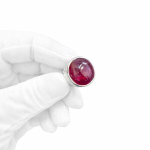 Może przedstawiać: Pierścionek z dużym, okrągłym kamieniem szlachetnym w kolorze rubinu, osadzonym w teksturowanej srebrnej oprawie. Pierścionek jest trzymany przez dłoń w rękawicy na białym tle. Kamień szlachetny ma głęboki czerwony kolor.