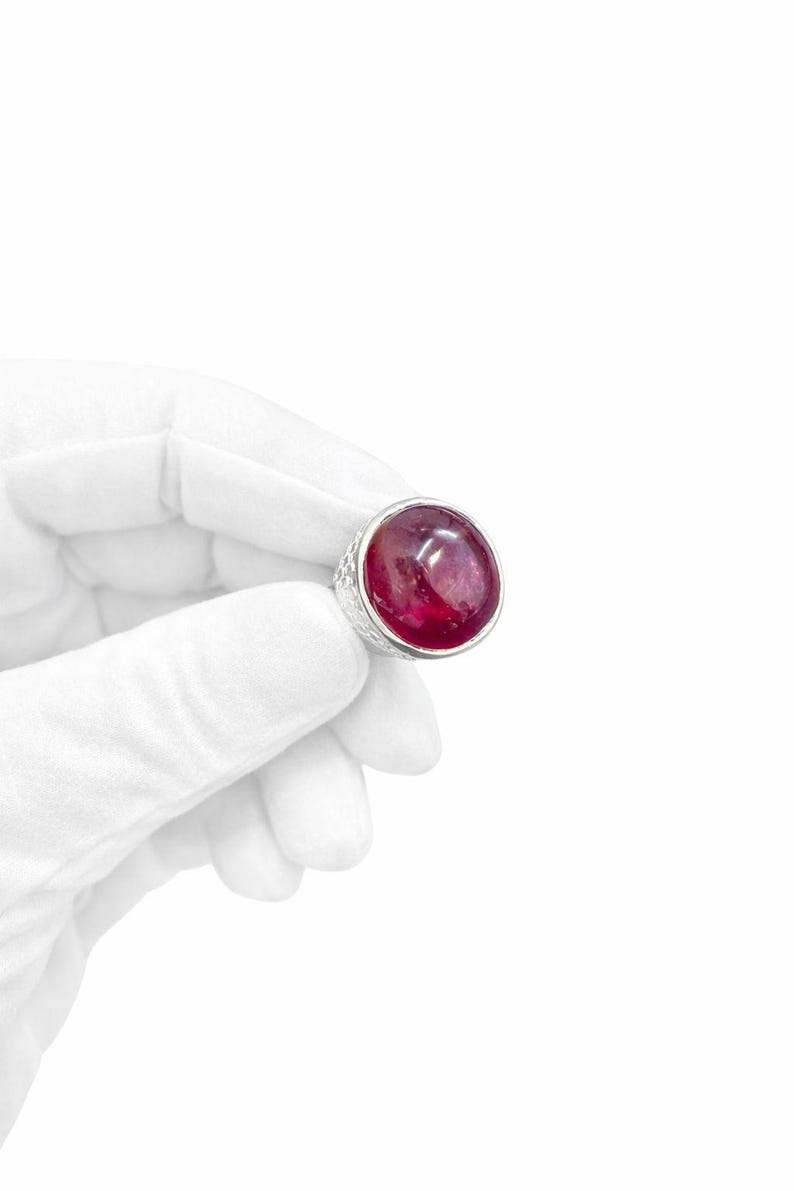 Może przedstawiać: Pierścionek z dużym, okrągłym kamieniem szlachetnym w kolorze rubinu, osadzonym w teksturowanej srebrnej oprawie. Pierścionek jest trzymany przez dłoń w rękawicy na białym tle. Kamień szlachetny ma głęboki czerwony kolor.