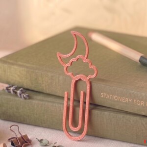 Puede incluir: Un clip de papel de color coral en forma de luna creciente y nube. El clip de papel está encima de dos libros de color verde oliva. Un lápiz descansa sobre los libros. El texto "STATIONERY FOR PEACE" es visible en el libro.