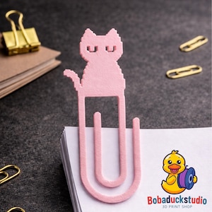 May include: A pink cat-shaped paper clip sits atop a stack of white paper. The cat design is pixelated. Other office supplies, including a gold binder clip and paper clips, are visible in the background. The logo "Bobaduckstudio 3D PRINT SHOP" is in the bottom right corner.