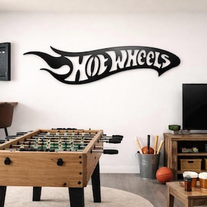 Pode incluir: Arte de parede com o logotipo "Hot Wheels" em metal preto. A placa tem um design de chama e é montada em uma parede branca. Uma mesa de pebolim, um dardo e uma TV também estão na sala.
