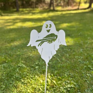 Op de afbeelding: Een witte, geestvormige tuinpaal van metaal met een uitsparing van het Buffalo Bills-logo in het midden. De geest heeft een lachend gezicht met twee ronde ogen en een open mond, gemonteerd op een dunne metalen staaf voor in de grond.