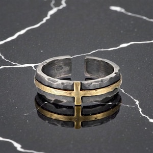 Könnte beinhalten: Verstellbarer Ring in Silberoptik mit gehämmertem Muster und einem goldfarbenen Kreuzdesign. Der Ring hat ein geteiltes Band und ein goldenes Band, das sich um die Mitte wickelt. Der Ring wird auf einer schwarz-weißen Marmoroberfläche präsentiert.