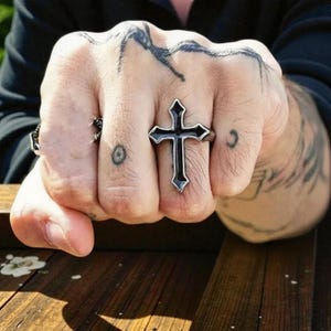 Puede incluir: Un anillo de cruz negra con borde plateado es el punto focal de esta imagen. El anillo se lleva en una mano con tatuajes. El diseño de la cruz es una pieza audaz, un símbolo de fe y estilo.