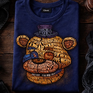 May include: Navy blue t-shirt featuring a graphic of Freddy Fazbear's face, composed of words and phrases. The design includes the text "Freddy," "Five Nights," and other related terms. The shirt has a classic fit.