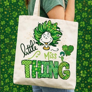 May include: A cream-colored tote bag featuring a whimsical green-haired character with a bow, the words "Little Miss Thing," and a heart-shaped shamrock design. The text and graphics are in shades of green.